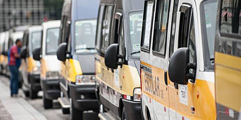 Mais de oito veículos de transporte escolar enfileirados, durante o dia. Ao fundo, um homemn conversa fora do veículo, voltado para a janela de um deles. 