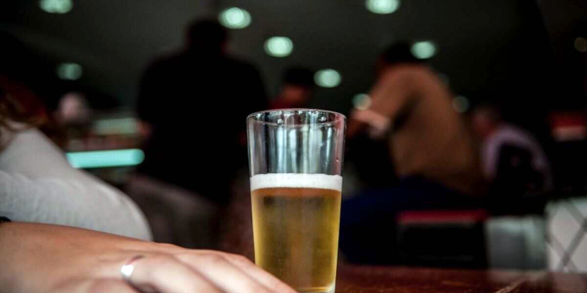 Um copo de cerveja em cima da mesa. Ao lado uma mão de mulher com unhas pintadas de vermelho . A iluminação do teto indica que é um bar ou uma  boate 
