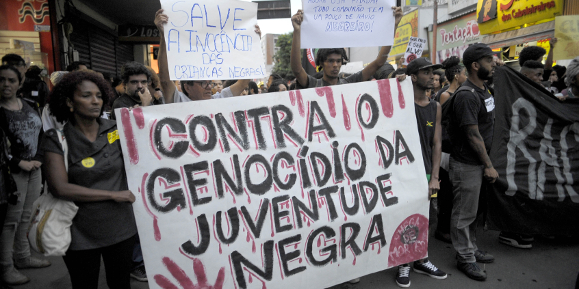 Em rua movimentada, mais de vinte pessoas em passeata seguram cartazes, entre eles dois com os dizeres "Contra o genocídio da juventide negra" e "Salve a inocência das crianças negras". 
