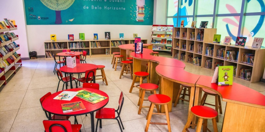 Loca para leitura infantil, com mesa e bancada para diferentes faixas etárias e prateleiras com livros diversos. Ao fundo, parede pintada de verde escuro com ilustração de duas crianças lendo e os seguintes dizeres ao meio: "Biblioteca Pública Infantil e Juvenil de Belo Horizonte".