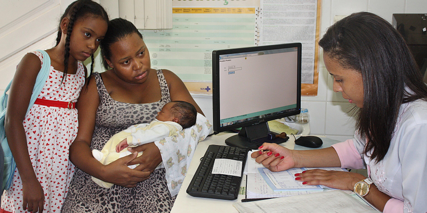 Mulher sentada com filho recém nascido no colo e filha de pé, ao lado, é atendida por médica que preenche um formulário, sentada à frete de mesa com computador. 