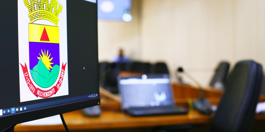Tela de computador mostra brasão de BH em primeiro plano. Ao fundo uma imagem desfocada de outro computador e uma cadeira vazia 