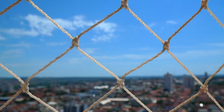 Janela com rede de proteção e paisagem urbana ao fundo, com árvores, casas e céu azul. 