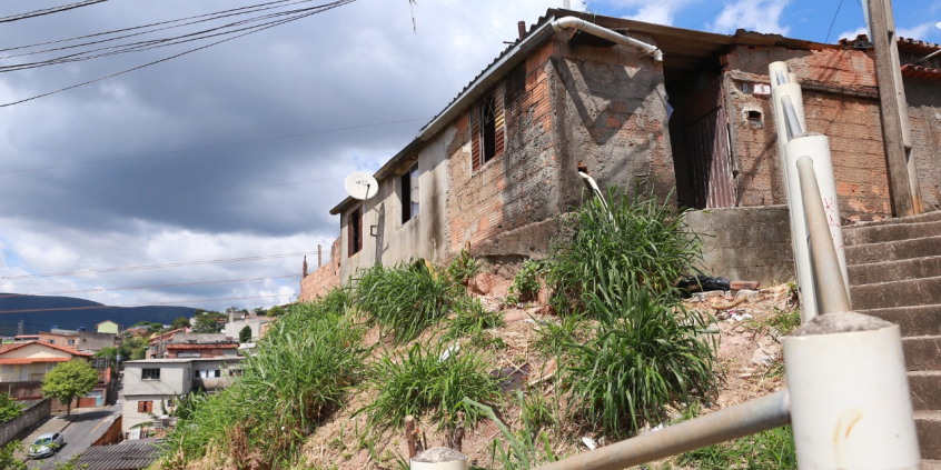 Casebre com tijolos aparentes e telhado de zinco no alto de um barranco com mato alto