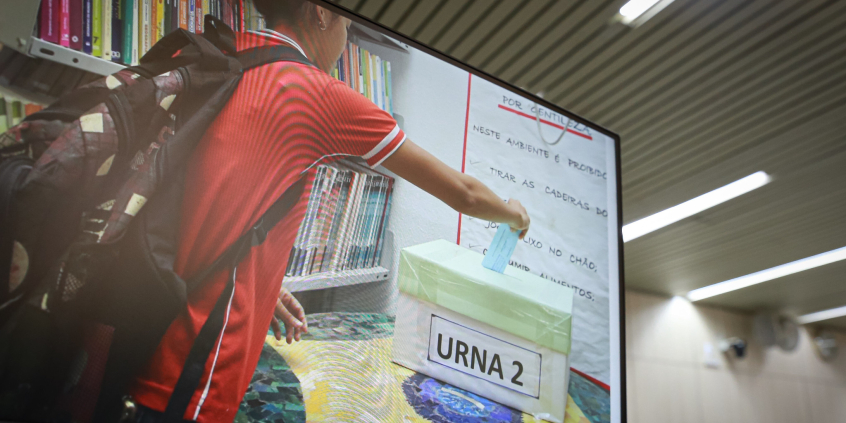 imagem na tela do computador mostra criança ,vestida com camisa vermelha e usando mochila preta nas costas, votando em uma urna  de papelão. Ao fundo, uma estante cheia de livros e um quadro branco nos remete á ideia de sala de aula 