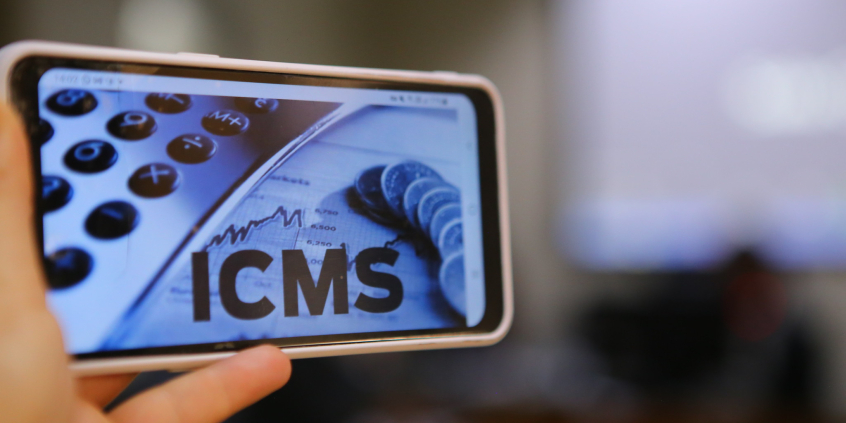 A tela do celular em primeiro plano mostra as teclas de uma calculadora e a sigla ICMS ao lado de algumas moedas