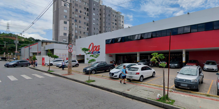 Rua em frente a supermercado, com carros estacionados e faixa para pedestres, durante o dia.