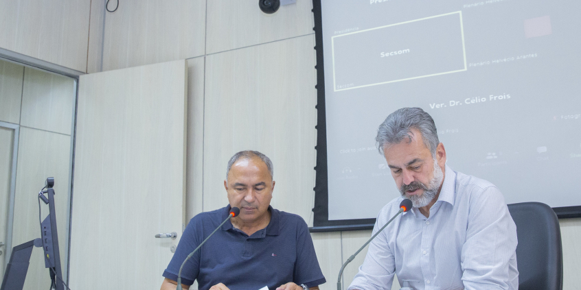 dois vereadores, um deles com um computador à frente, sentados à mesa, em reunião presencial. 