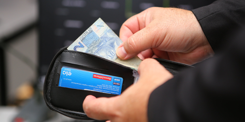 Um homem tira uma nota de dois reais de uma carteira preta. Ele veste um paletó preto