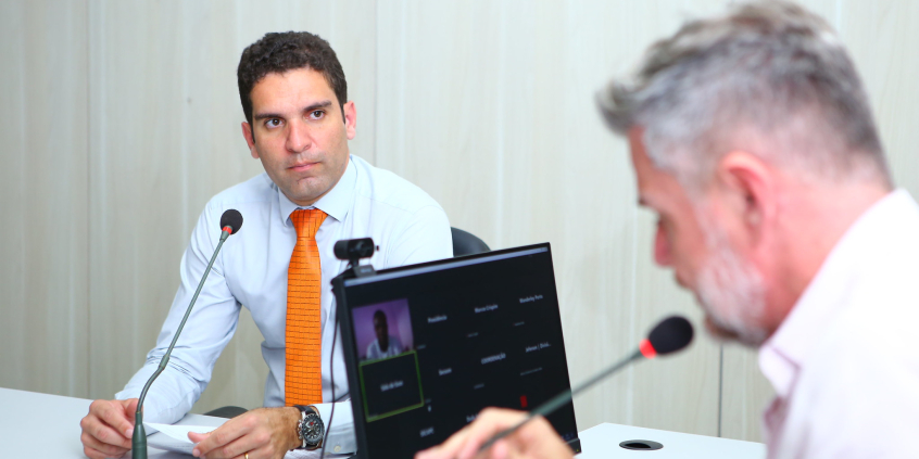 Dois vereadores sentados em reunião presencial e um deles exibido em tela de computador, participando de maneira remota. 