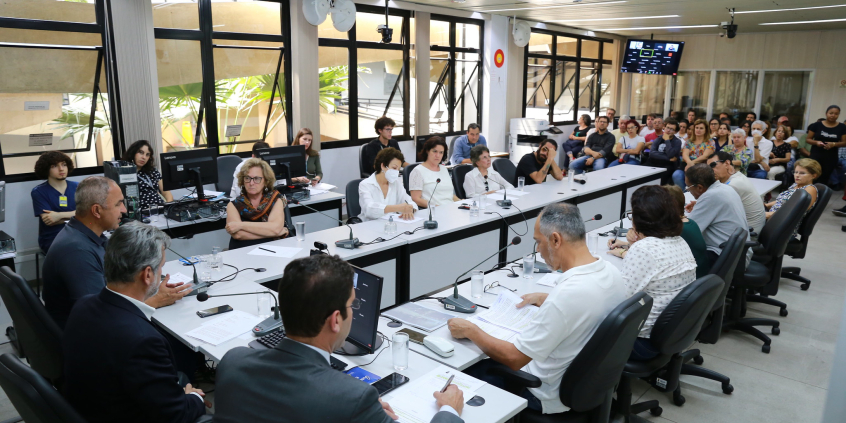 três vereadores à esquerda, dez participantes ao logo da mesa em formato de "u" e mais de vinte cidadãos acompanham audiência pública