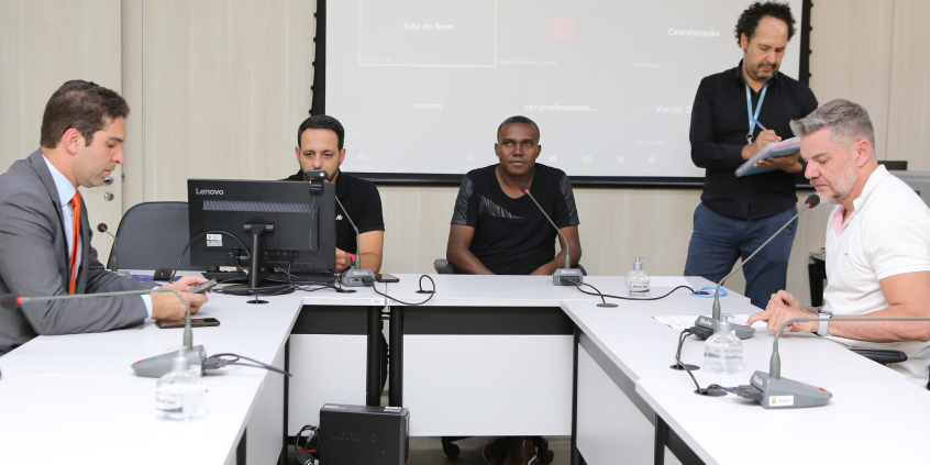 Quatro vereadores sentados à mesa, em reunião presencial, acompanhados de Assessor da Câmara, de pé.
