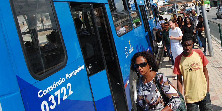 Cerca de 14 pessoas estão próximas ao ponto de ônibus, durante o dia. Três delas estão embarcando em ônibus coletivo..