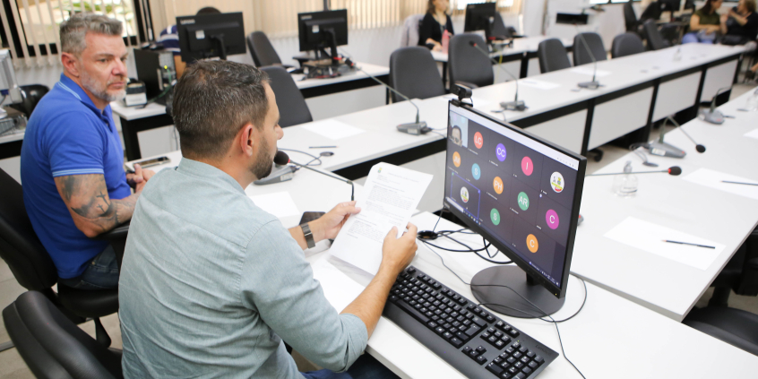Dois vereadores sentados à mesa em reunião presencial, um deles tem tela de computador à frente na qual outros vereadores participam de maneira remota.