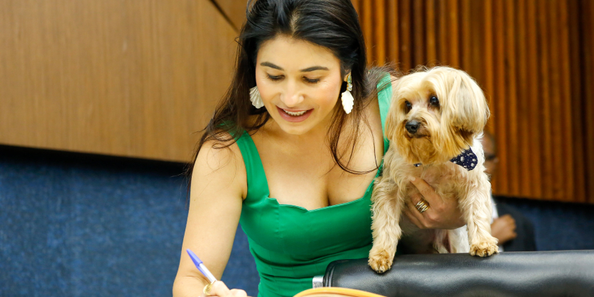 Mulher assina livro enquanto segura cão no braço esquerdo.