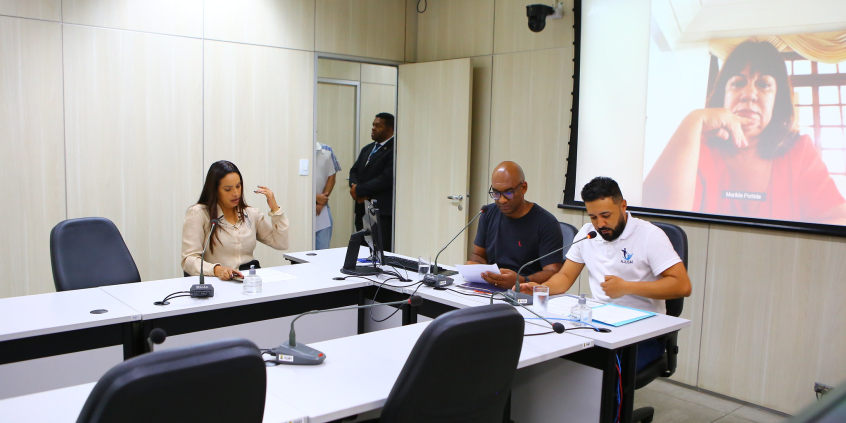 Dois vereadores e uma vereadora sentados à mesa, reunidos. No fundo da sala, telão exibe a imagem de outra vereadora, que participa da reunião de maneira remota.