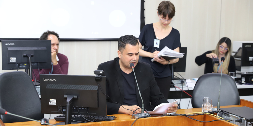 Vereador conduz reunião, entado à mesa, do lado de uma tela de computador.