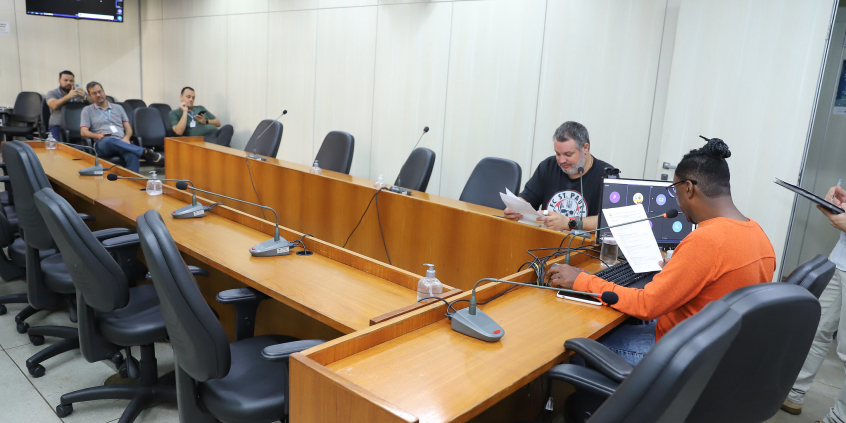 Dois parlamentares sentados à mesa, em reunião, com três homens assistindo , ao fundo.