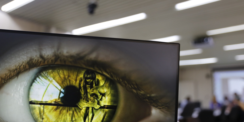 imagem de um olho verde na tela do computador. é possível ver o reflexo de uma mulher sentada de cabeiça baixa diante do punho cerrado de um homem