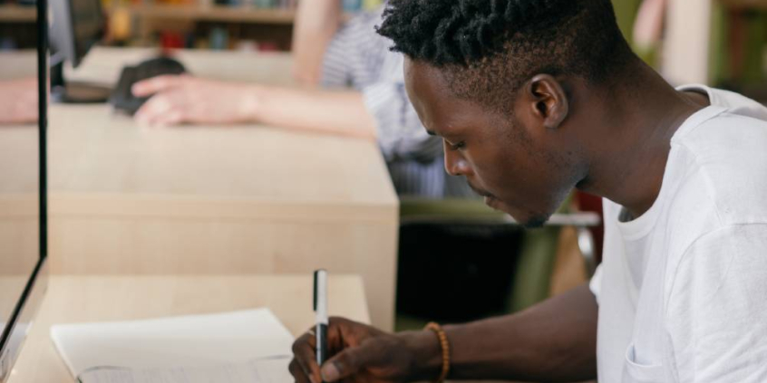 Jovem negro faz anotações em caderno, sentado à frente de computador.
