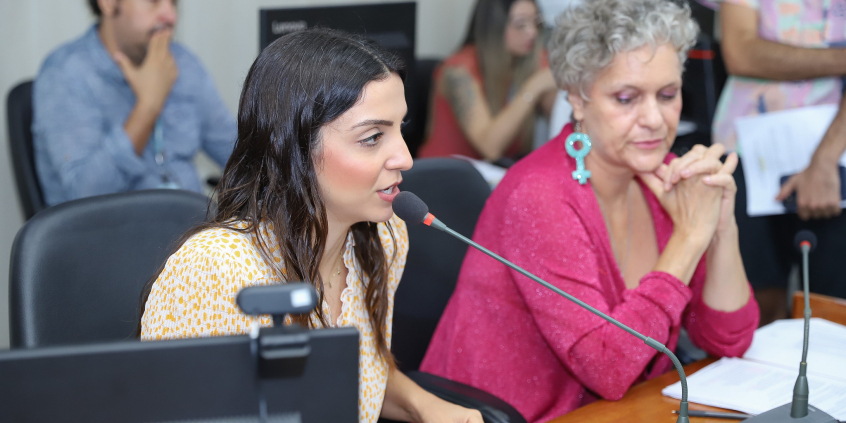 Imagem das vereadoras Marcela Trópia (Novo) e Cida Falabella (Psol) sentadas à mesa , durante reunião da Comissão