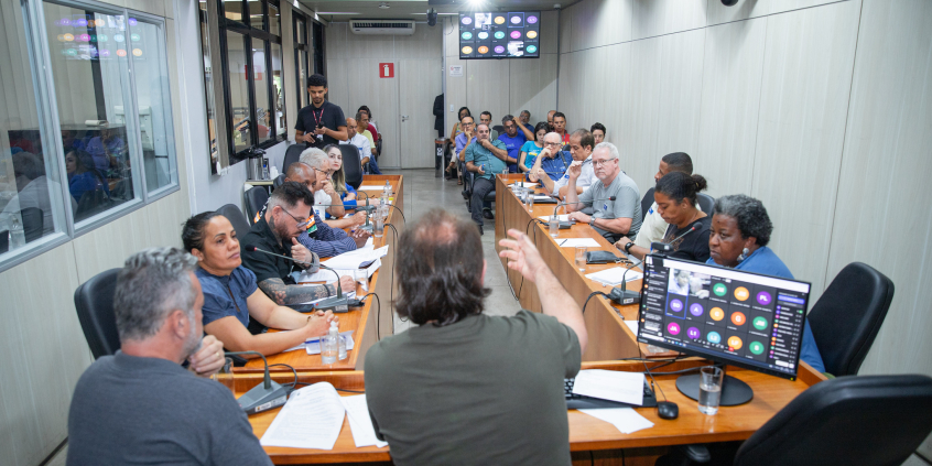Dois vereadores e uma deputada estadual reunidos à mesa com dez cidadãos, com maid e dez pessoas sentadas em cadeiras ao fundo da sala.
