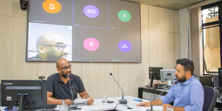Dois vereadores participam de reunião, sentados à mesa.