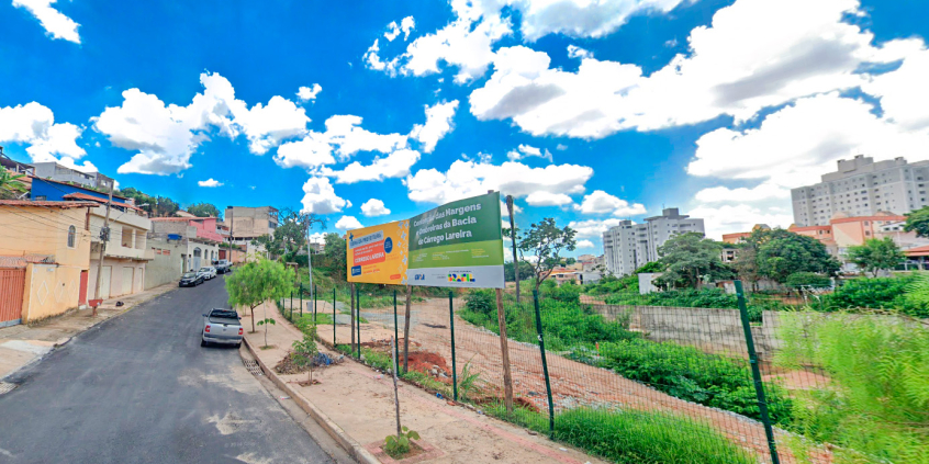 Rua urbanizada com placa de obra no córrego Lareira, à direita.