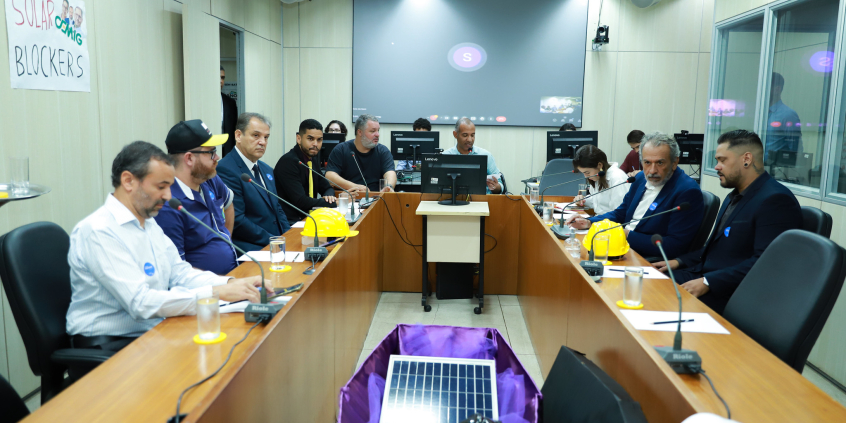 Nove pessoas reunidas, sentadas à mesa. No centro, um caixão uma célula de produção de energia solar acompanhada por capacetes de construção.
