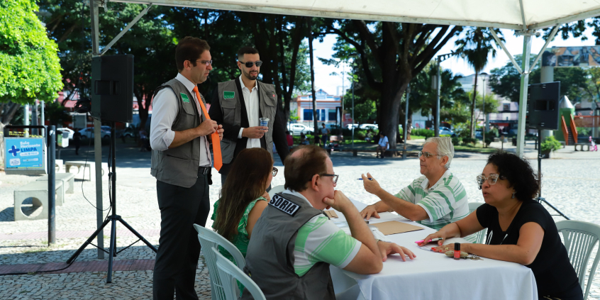 Um homem e uma mulher se consultam com profissionais da Ouvidoria Itinerante sentados à mesa, em praça, durante o dia, acompanhados pelo ouvidor e outro servidor da Ouvidoria. 