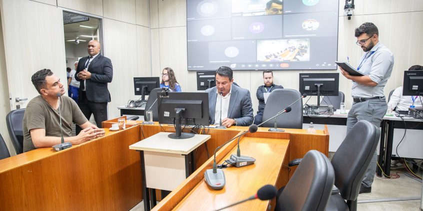 Fernando Luiz e Irlan Melo estão assentados no Plenário Camil Caram. O primeiro vereador fala ao microfone. Atrás dele, o telão mostra os outros parlamentares presentes em chamada de vídeo.