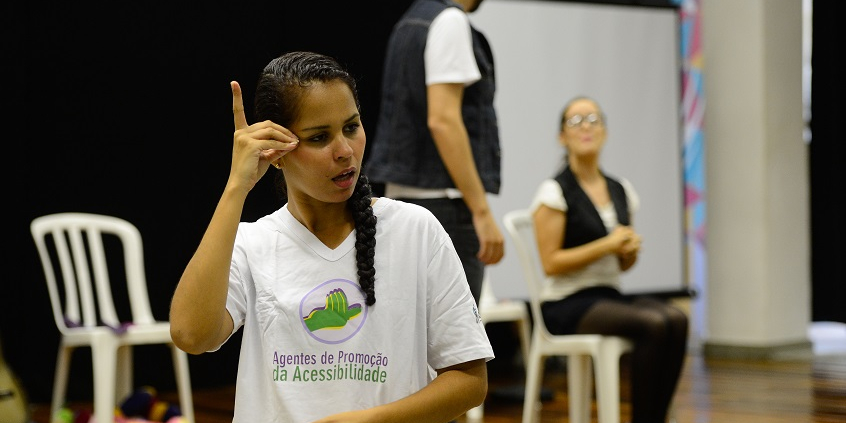 Mulher com camisa branca escrito "Agentes de promoção da acessbilidade", faz um sinal em Libras