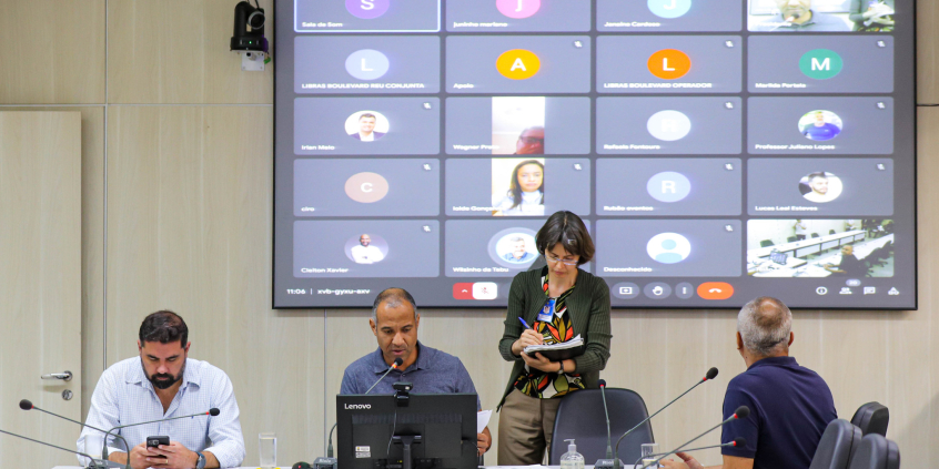 Vereadores estão sentados no plenário Helvécio Arantes. O telão atrás deles mostra a videochamada com parlamentares presentes virtualmente.