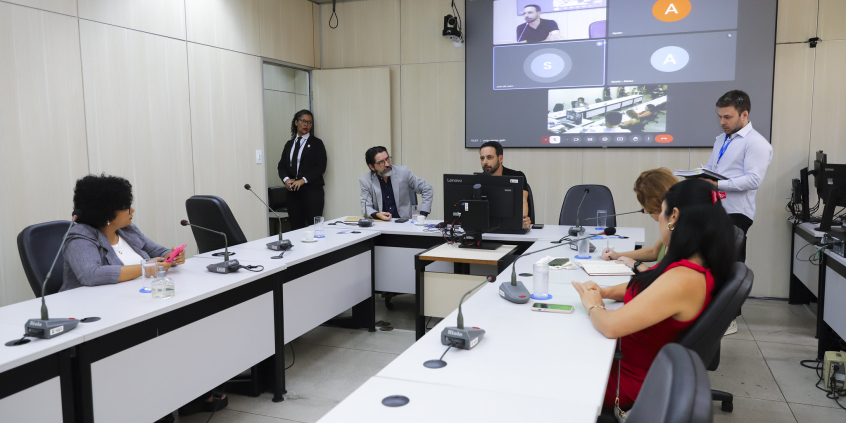 Vereadores no Plenário Helvécio Arantes, sentados com microfones na mesa à frente deles, e um telão atrás.