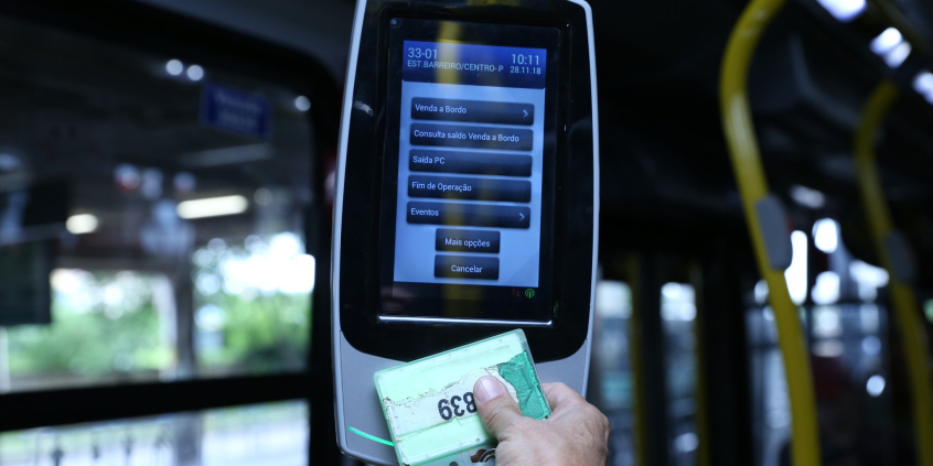 Na foto uma mão encosta um cartão na máquina de cobrança de passagem de um ônibus