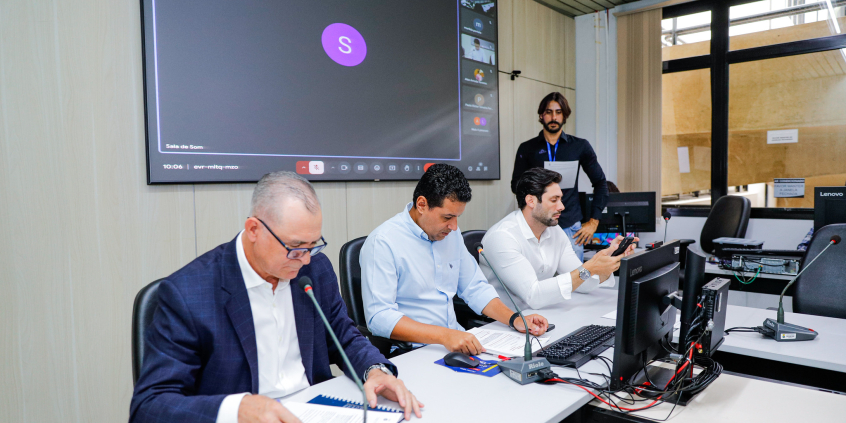 Vereadores estão sentados no plenário. O telão atrás deles mostra a videochamada com parlamentares presentes virtualmente.