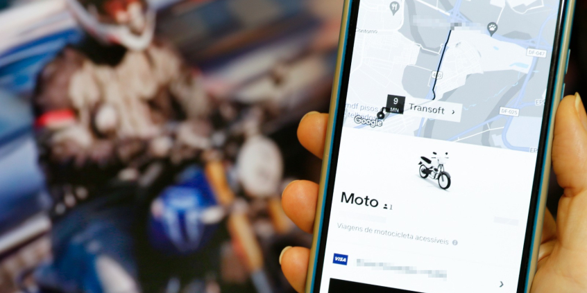 Em primeiro, plano, celular com aplicativo de corrida por moto. Em segundo plano, motociclista