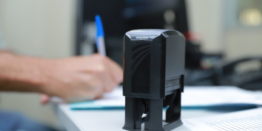 A foto mostra um carimbo em cima de uma mesa.  Atrás dele, uma mão escreve à caneta.