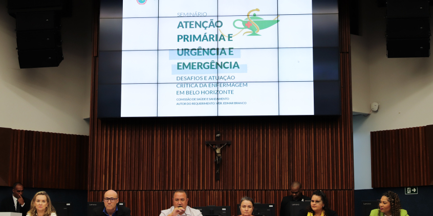 Membros da mesa de debate do seminário de enfermagem estão assentados no Plenário Amintas de Barros; acima deles está um telão com um slide