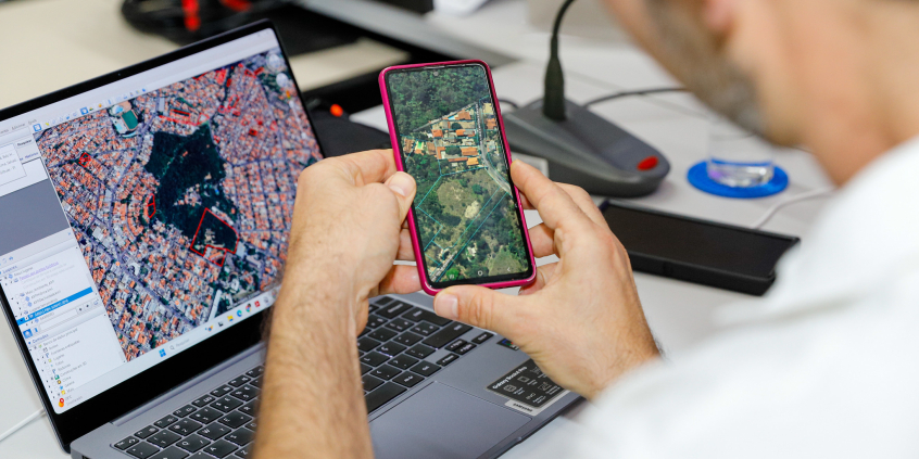 homem com o celular na mãe e o computador à frente, ambos com imagem geográfica de satélite