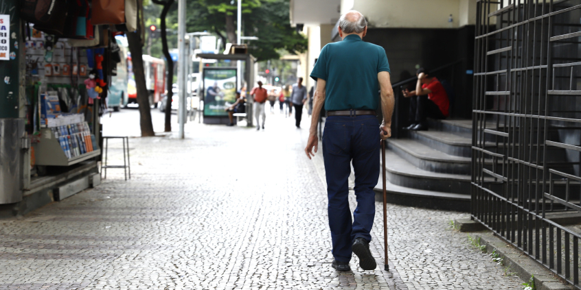 Idoso visto de costas caminha em rua de BH com bengala