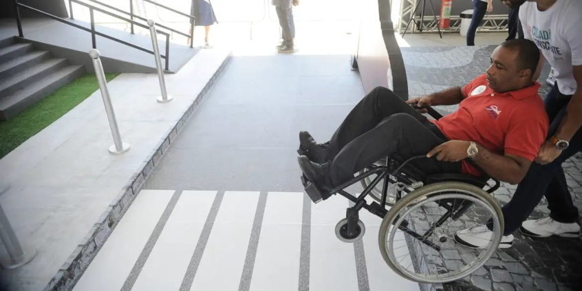 homem cadeirante recebe ajuda de outro homem para subir rampa