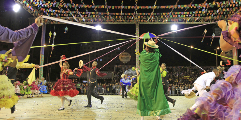 Grupo junino dançando com bandeiras ao fundo no Arraial de Belo Horizonte