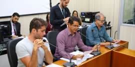 Três integrantes da comissão estão sentados à Mesa do Plenário Camil Caram; um servidor de pé e três assentados atrás deles assessoram e registram as ocorrências da reunião.