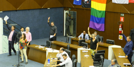 Alguns vereadores estão assentados, outros, de pé, se manifestam durante a reunião; atrás, vêm-se bandeiras pendendo da galeris