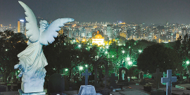 Vista noturna de lápides e esculturas no Cemitério do Bonfim. Ao fundo, luzes da cidade. Vista panorâmica da cidade