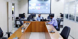 Parlamentares compõem mesa de reunião. Telão ao fundo em videoconferência com os demais membros e convidados da CPI