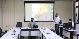 Parlamentar compõe mesa de reunião. Telão ao fundo em videoconferência com os demais membros da comissão