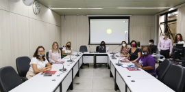 Sete mulheres, todas de máscara, sentadas ao redor de uma mesa em formato de "u", em uma sala. Ao fundo, um telão e à direita, um homem e uma mulher, de pé, e um homem ao fundo sentado diante de um computador, os três de mascara.