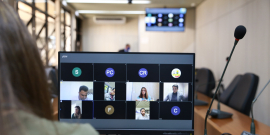 Monitor de computador exibe vereadores em videoconferência. Ao fundo, cadeiras vazias do Plenário Camil Caram